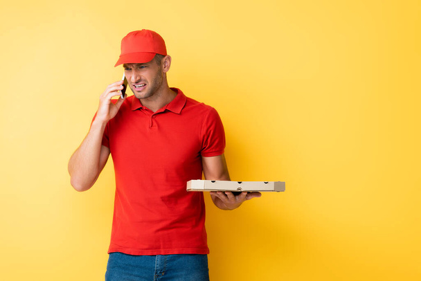 displeased delivery man in red cap holding box with tasty pizza and talking on smartphone on yellow - Fotoğraf, Görsel