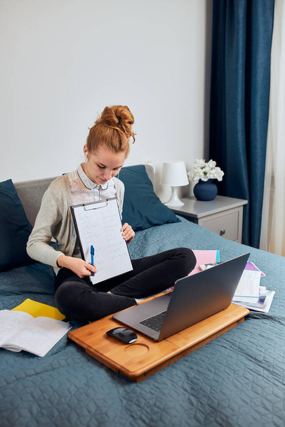 Young woman student having classes, learning online, watching lesson remotely, listening to professor, talking with classmates on video call from home during quarantine. Young girl using laptop, headphones, books, manuals sitting on bed - Foto, Imagem