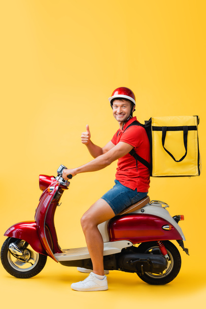 happy delivery man in helmet and backpack riding red scooter while showing thumb up on yellow - Φωτογραφία, εικόνα