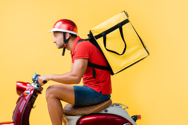 side view of delivery man with backpack riding red scooter on yellow - Foto, Imagem