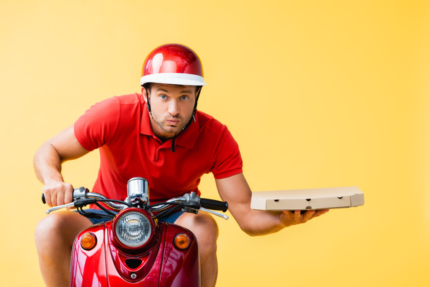 концентрований чоловік доставки в шолом їде червоним скутером і тримає коробку для піци ізольовано на жовтому
 - Фото, зображення