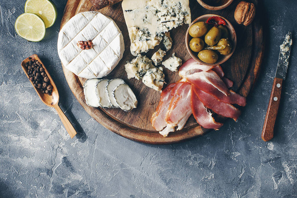 Set of cheese, hamon, honey, green olives, nuts and spices on rustic wooden board, variety of snacks on dark stone table, top view - Foto, afbeelding