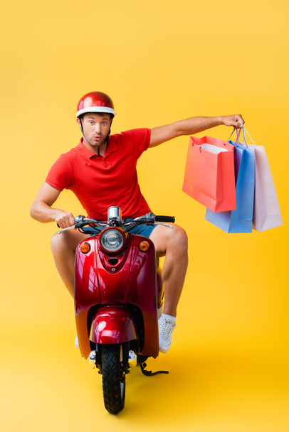 surprised delivery man in helmet riding scooter and holding shopping bags on yellow  - Fotografie, Obrázek