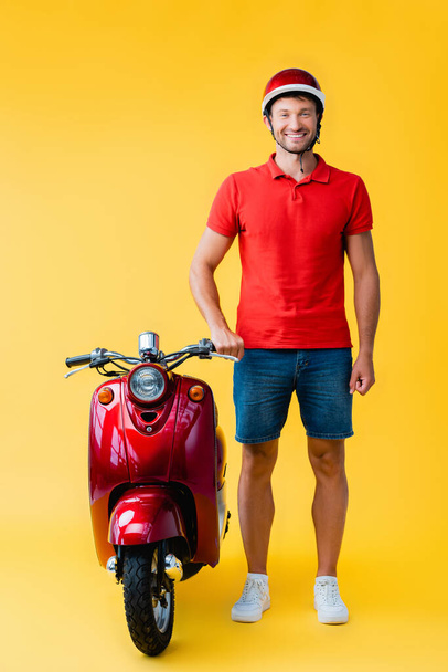 full length of happy man in helmet standing near red scooter on yellow - Foto, Imagem