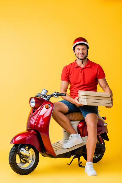 hombre de entrega feliz en casco sosteniendo cajas de pizza mientras está sentado en scooter en amarillo - Foto, Imagen