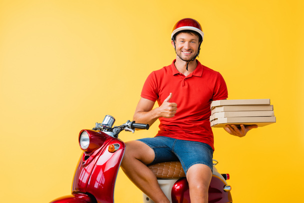 glücklicher Zusteller im Helm, der Pizzakartons hält, während er den Daumen in der Nähe von Roller auf gelb zeigt - Foto, Bild