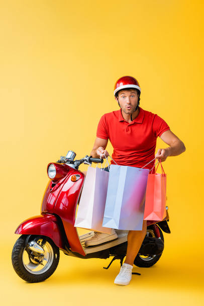geschokt levering man in helm zittend op scooter en het houden van boodschappentassen op geel  - Foto, afbeelding