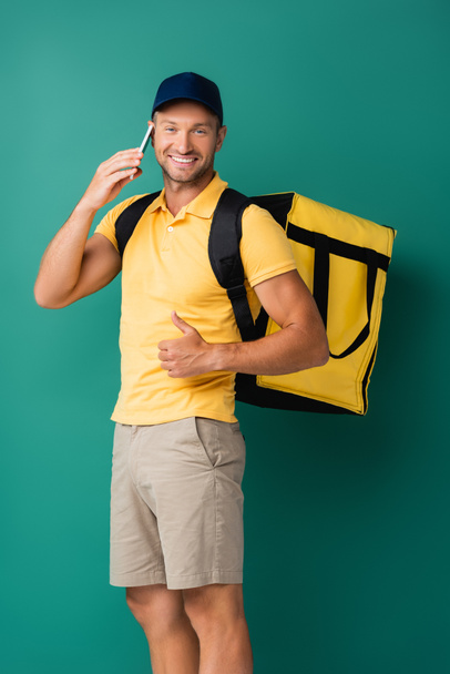 cheerful delivery man carrying yellow backpack and talking on smartphone on blue - Φωτογραφία, εικόνα