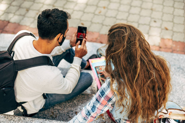 Takanäkymä kaksi nuorta opiskelijaa etsii matkapuhelimeen istuu portaat college kampuksen ulkona. Nuori nainen ja ulkona istuva mies käyvät online-keskusteluja älypuhelimella. - Valokuva, kuva