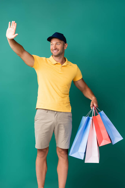 cheerful delivery man in cap carrying shopping bags and waving hand on blue - Photo, Image