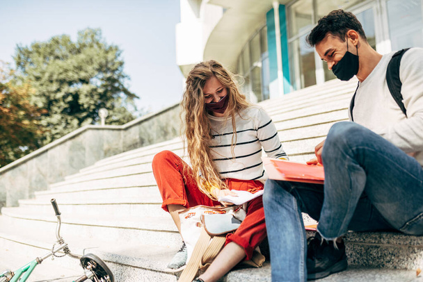 Outdoor image of a young woman and a man sitting on the stairs of the university for team work. Two young students in masks learning together sitting on the stairs of the college campus outside. - Fotografie, Obrázek