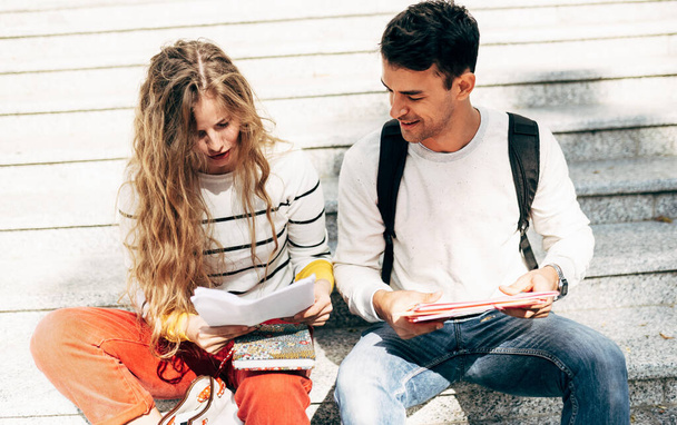 Horizontal image of two young students sitting on the stairs and learning together at the university outside. Two colleagues preparing for exams at the college campus outdoors. - Valokuva, kuva