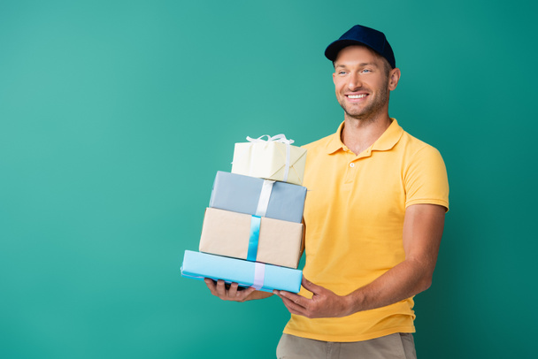 positive delivery man in cap holding wrapped presents on blue - Foto, afbeelding