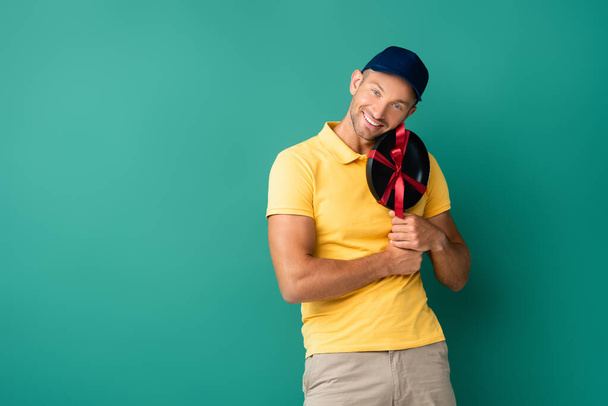 cheerful delivery man in cap holding frying pan with ribbon on blue  - Photo, Image