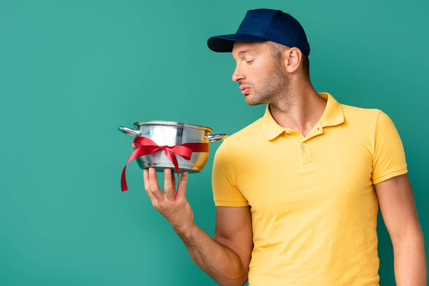 delivery man in cap looking at saucepan with ribbon on blue  - Photo, Image