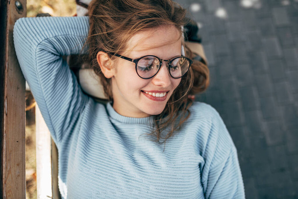 Gorgeous female in blue sweater, glasses, smiling with closed eyes, take a rest on the bench outside. Happy woman takes a break after college outside. - Foto, Imagen