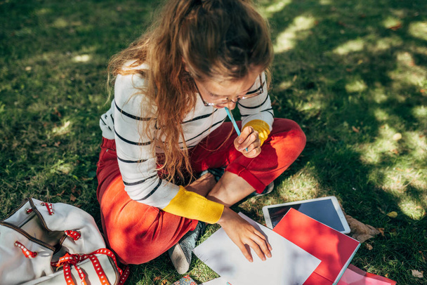Top view of a student female in casual outfit, glasses, sitting on the green grass at the college campus, and studying with lots of books outdoors. Young woman learning and making notes in the park. - Valokuva, kuva