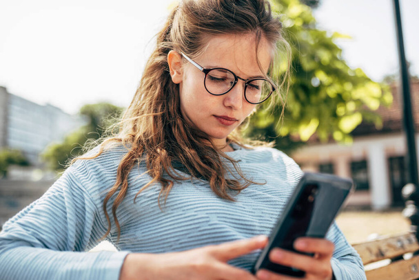 Pretty female in blue sweater and glasses, sitting on the bench messaging on mobile phone outdoors. A beautiful young woman takes a rest during learning online on a smartphone outside. - 写真・画像
