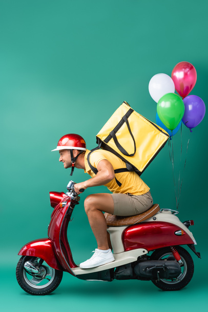 side view of focused delivery man with backpack riding scooter with balloons on blue  - Zdjęcie, obraz