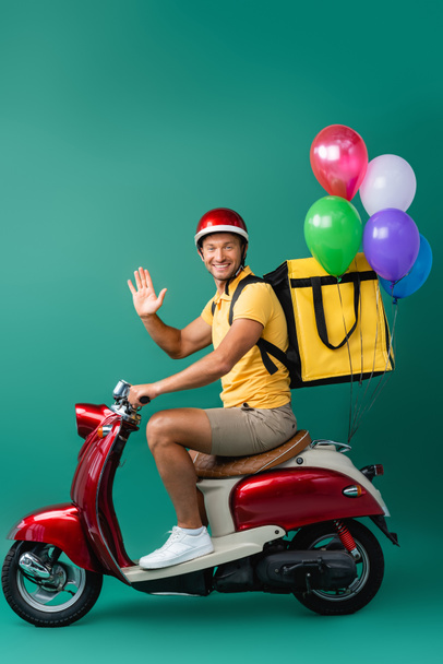 happy delivery man with backpack riding scooter with balloons and waving hand on blue  - Fotó, kép