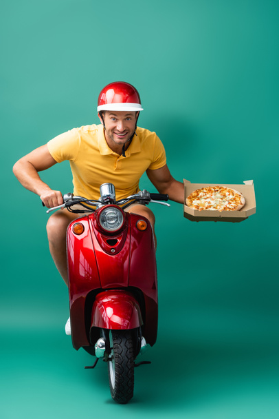 veselé dodávky muž v helmě na koni skútr při držení chutné pizzy v krabici na modré - Fotografie, Obrázek