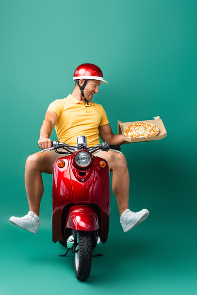 excited delivery man in helmet riding scooter while holding tasty pizza in box on blue - 写真・画像