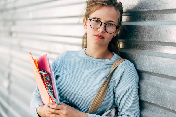 Horizontal outdoor image of a pretty female student carrying lots of books after a day in the college. Beautiful young woman wearing a blue sweater and glasses posing with books outside on the street. - Photo, image