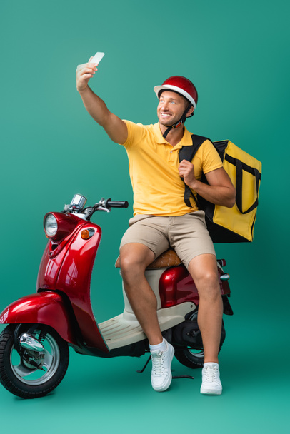 alegre entregar hombre con mochila tomando selfie cerca de scooter en azul - Foto, imagen