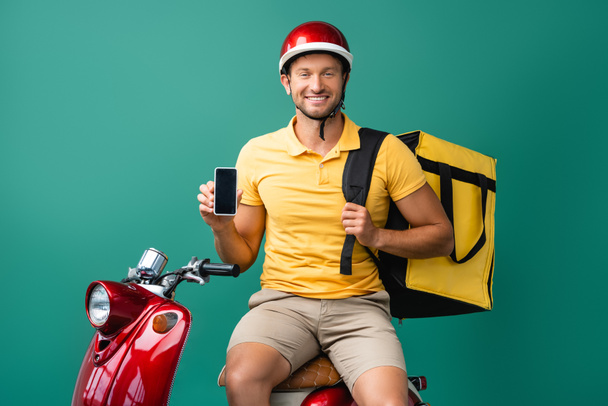 happy delivery man with backpack holding smartphone with blank screen near scooter on blue - Foto, imagen