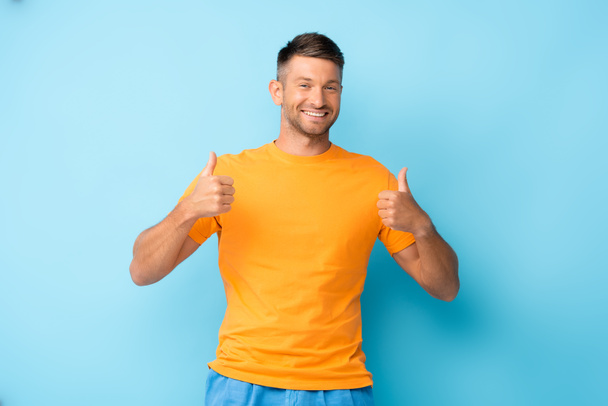 Ευτυχισμένος άντρας με κίτρινο μπλουζάκι που δείχνει τους αντίχειρες πάνω στο μπλε - Φωτογραφία, εικόνα