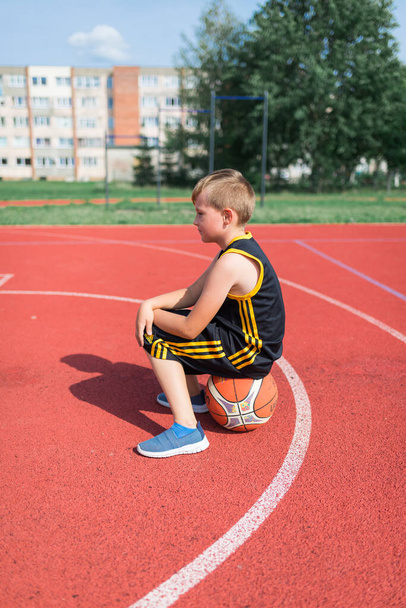Vista lateral del niño de la escuela primaria triste sentado en el baloncesto - Foto, Imagen