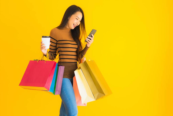 Krásná mladá asijská žena používat chytrý mobilní telefon nebo mobil s šálkem kávy a barva nákupní taška na žlutém izolovaném pozadí - Fotografie, Obrázek