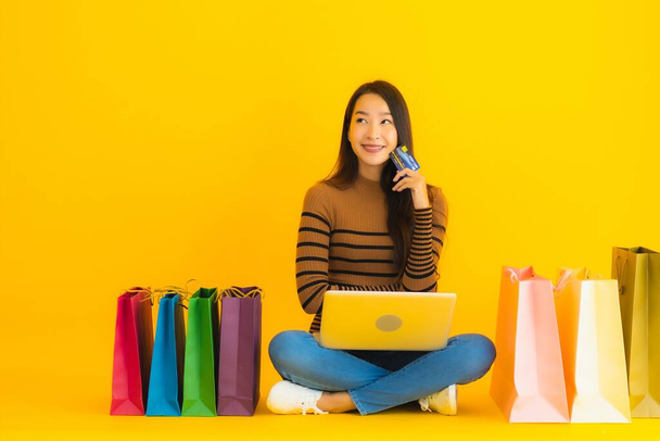 Mooi portret jonge aziatische vrouw zitten op de vloer met laptop of computer gebruik credit card voor online winkelen rond boodschappentas op gele achtergrond - Foto, afbeelding