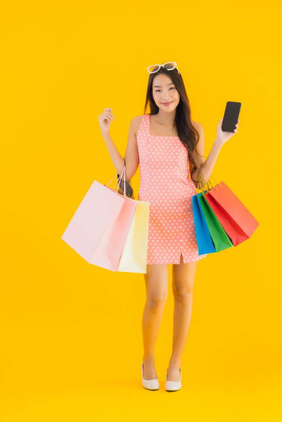 Портрет красивої молодої азіатки з барвистою сумкою для покупок зі смартфоном та кредитною карткою на жовтому ізольованому фоні
 - Фото, зображення