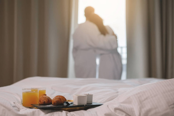 Breakfast tray is on a white bed. A man and a woman in robes hugging at the window in the background. Selective focus. - Photo, Image