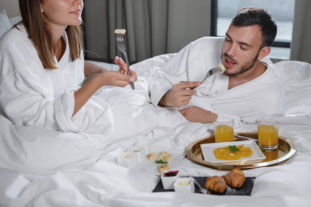 Un hombre barbudo y una mujer con batas blancas desayunan en la cama. Enfoque selectivo. - Foto, Imagen