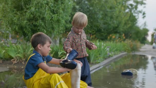 buiten kinderen, jongens in overalls spelen in plas door de weg na regen en gieten water uit laarzen na plezier - Video