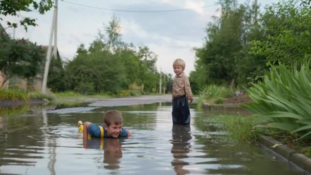 dzieciństwo, szczęśliwy chłopiec z dobrym zdrowiem, wraz z młodszym bratem, lubi gry i leży w kałuży w ciepły letni dzień po deszczu na drodze - Materiał filmowy, wideo