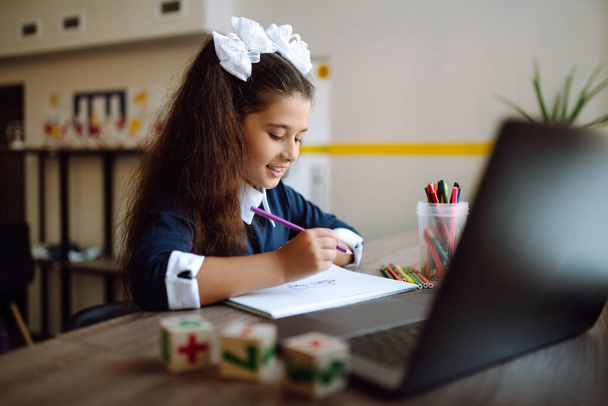 Beautiful school girl sitting at the table, draws and studying homework. School girl using laptop computer studying through online e-learning system.  Distance learning.  Covid-2019. - Photo, image