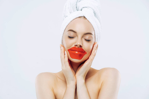 Cosmetology and skin care concept. Pretty woman with calm expression, closes eyes, applies lips patches, wears natural makeup, wrapped towel on head, stands bare shoulders indoor, white background - Фото, изображение