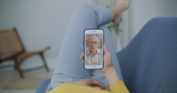 自宅の携帯電話の画面上でカンファレンスチャットオンラインアプリケーションを使用して、古い親の父や成熟した友人を呼び出す若い女性の娘のビデオの肩のビュー。ファミリービデオコンセプト - 映像、動画
