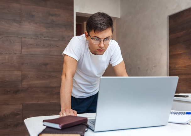 Concentrated man in glasses stands near a desk and looks at a laptop - Photo, image