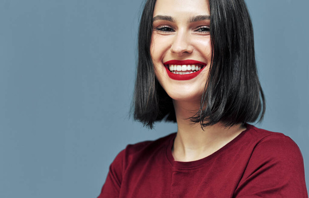 Closeup studio portrait of cheerful young woman with red lips smiling broadly with white teeth.  Brunette female model with makeup has joyful expression on blue background. - Foto, Bild