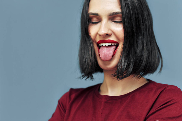 Studio portrait of playful young woman with red lips sticking tongue out. Brunette female model with makeup has happy and funny expression on blue background. - Photo, image