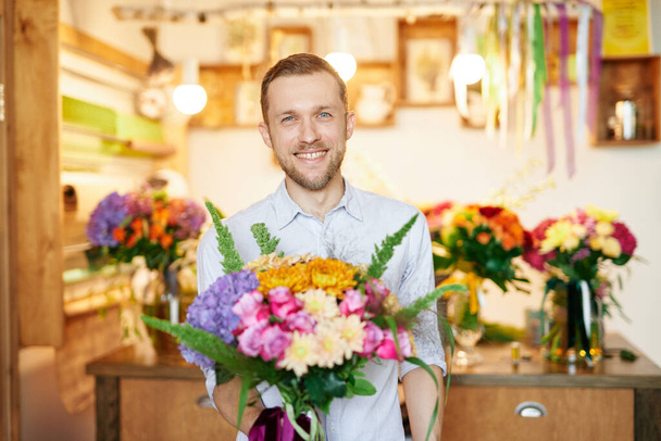 Jeune bel homme souriant avec bouquet dans la boutique de fleurs. Beau fleuriste homme en chemise tenant bouquet avec un large sourire. Concept d'entreprise. - Photo, image