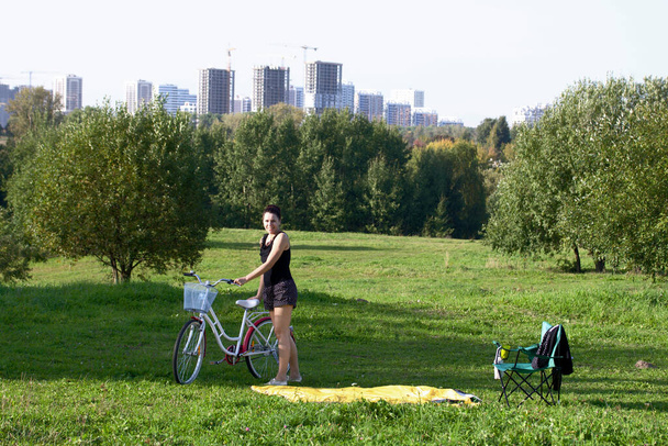 The girl is resting in the park. Stands, holds the bike in his hands. There is a camp chair nearby. - Foto, Imagem
