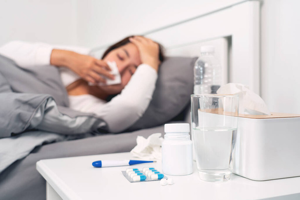 χάπι και ποτήρια νερό στο τραπέζι γυναίκα που πάσχουν από πονοκέφαλο, πυρετό και γρίπη που βρίσκεται στο κρεβάτι φόντο. - Φωτογραφία, εικόνα
