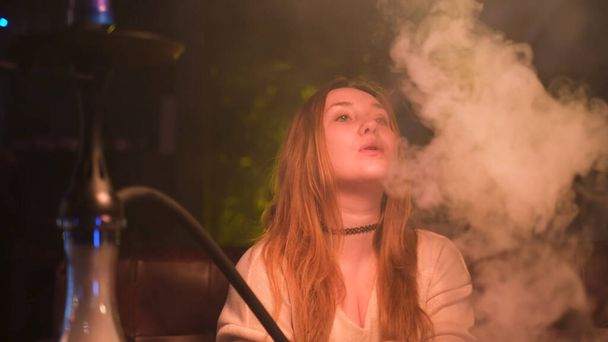 Retrato de cerca de una joven mujer de cabello castaño sexy fumando narguile sobre un fondo oscuro. Medios. Encantadora chica pensativa solitaria fumando shisha en el bar. - Foto, imagen