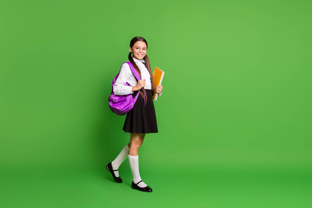 Photo portrait of schoolgirl going forward holding books in hand wearing violet backpack on shoulder isolated on vivid green colored background with copyspace - Photo, Image