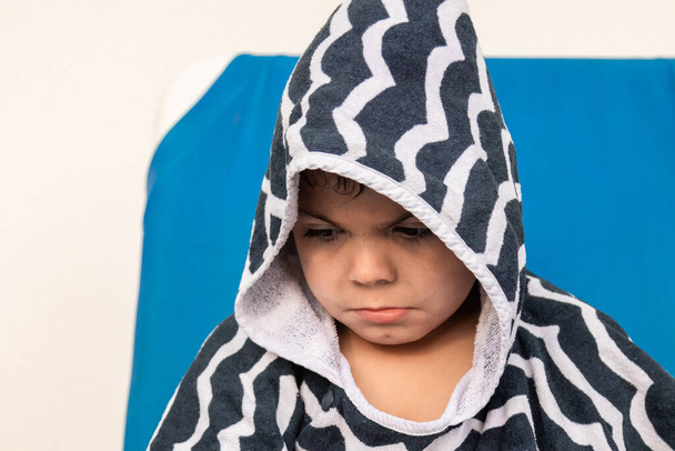 Der hispanische kaukasische Junge mit braunen Haaren ist wütend und traurig und trägt einen blau-grau gestreiften Bademantel mit Kapuze. Fotoporträt - Foto, Bild